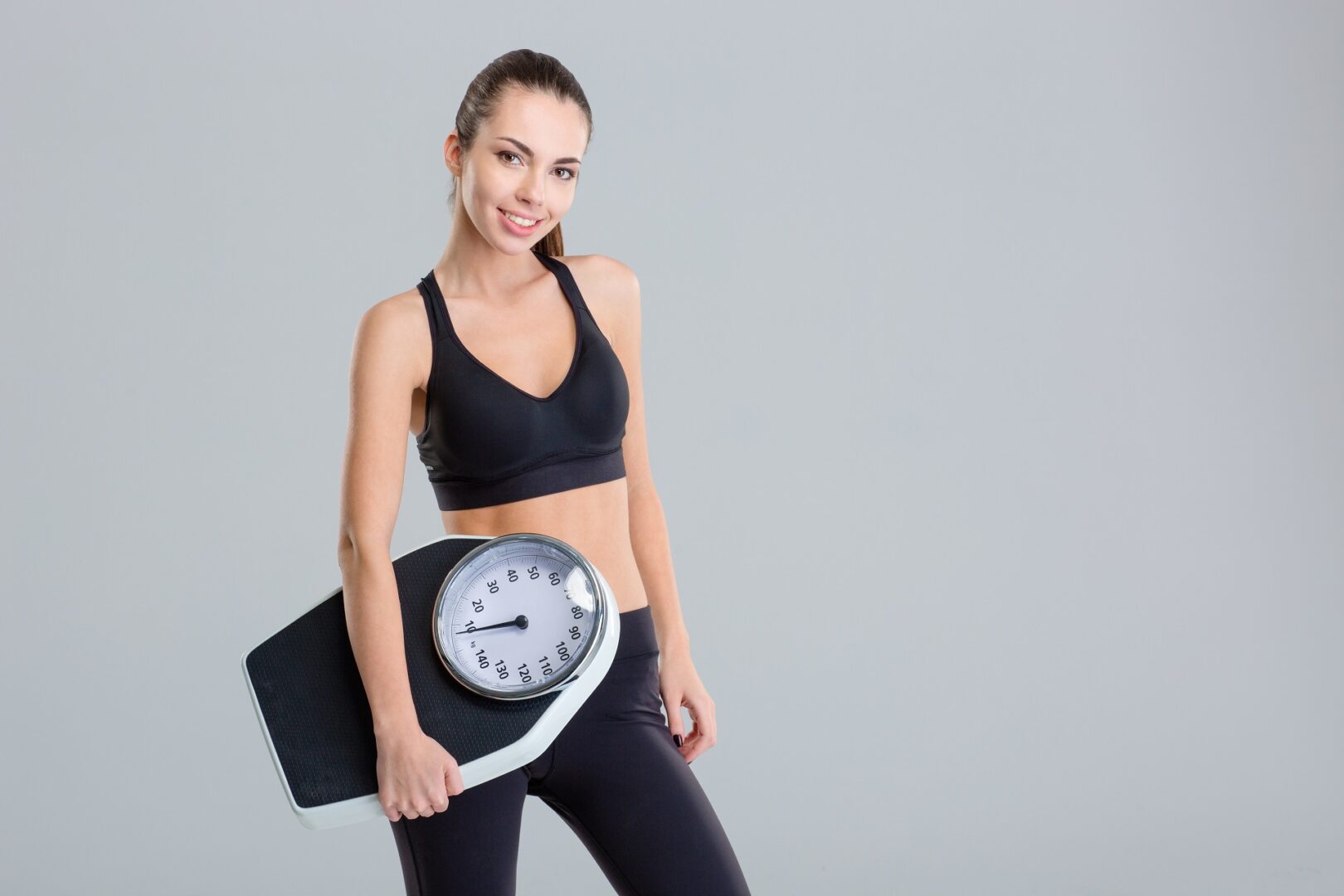 Beautiful smiling young fitness woman in tracksuit holding weigh scale over grey background